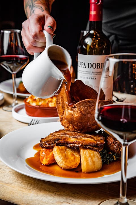 Gravy poured from a jug over a roast with Yorkshire pudding, a bottle of red wine behind